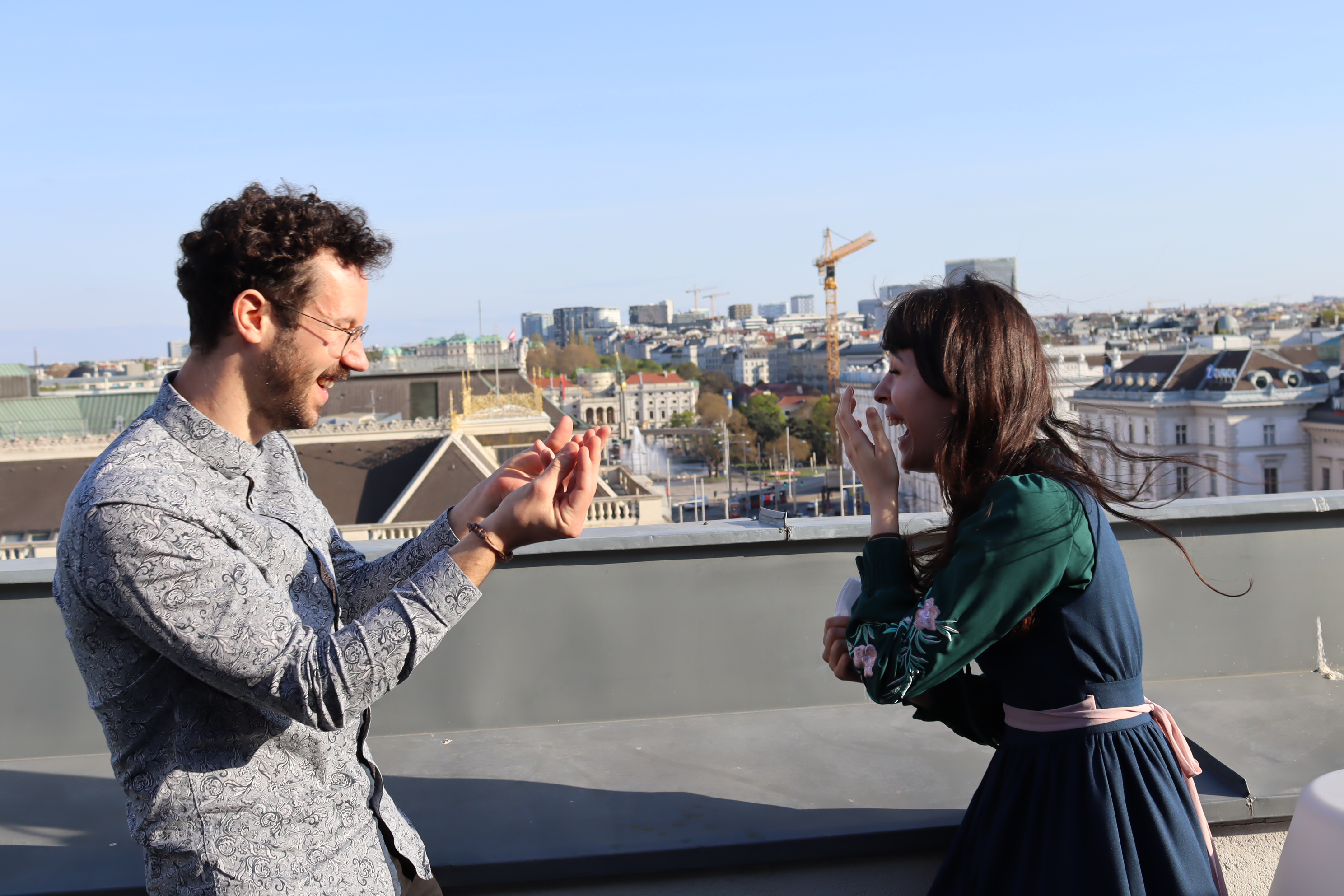 A man and a woman standing on a sunny day on a rooftop. He tries to explain something and she is laughing.